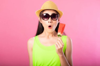 Yaz yeşil kıyafet şapka ve güneş gözlüğü üzerinde pembe arka plan izole sürpriz arıyor sopa taze karpuz holding ile mutlu bir çekici kadın portresi. Yaz vibe kavramı.