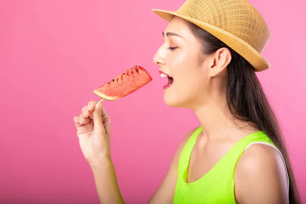 Portrait of a happy attractive woman in summer green outfit with hat biting fresh water melon on stick isolated over pink background. Summer vibe concept.