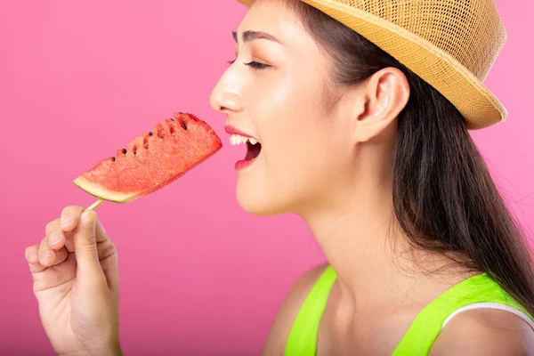 Portrait of a happy attractive woman in summer green outfit with hat biting fresh water melon on stick isolated over pink background. Summer vibe concept.