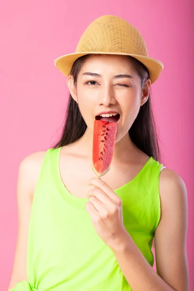 Portrait of a happy attractive woman in summer green outfit with hat biting fresh water melon on stick isolated over pink background. Summer vibe concept.