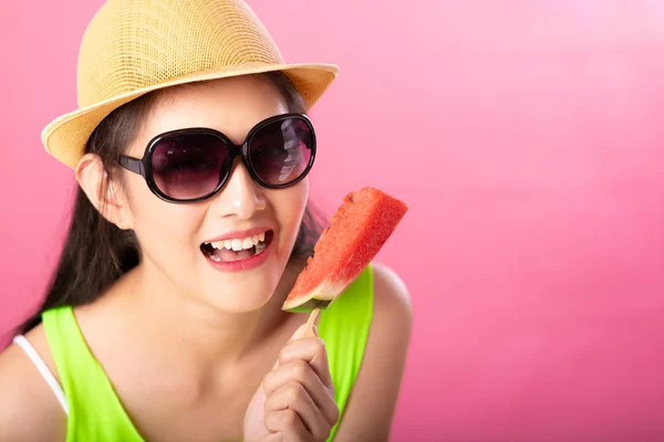 Portrait of a happy attractive woman in summer green outfit with hat and sunglasses holding fresh water melon on stick isolated over pink background. Summer vibe concept.