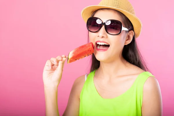 Portrait of a happy attractive woman in summer green outfit with hat and sunglasses biting fresh water melon on stick isolated over pink background. Summer vibe concept.