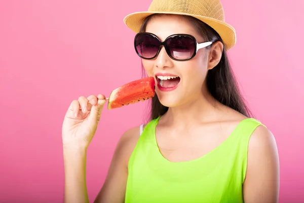 Portrait of a happy attractive woman in summer green outfit with hat and sunglasses biting fresh water melon on stick isolated over pink background. Summer vibe concept.