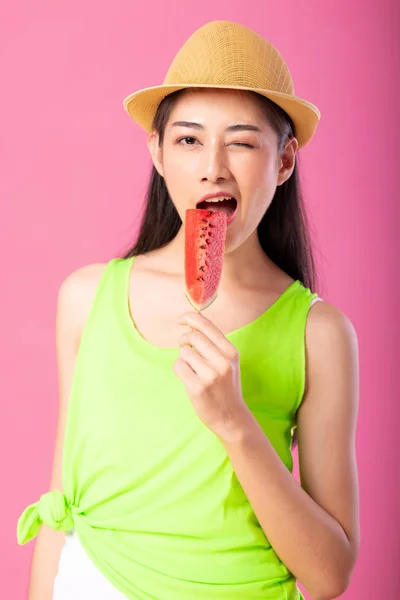 Portrait of a happy attractive woman in summer green outfit with hat biting fresh water melon on stick isolated over pink background. Summer vibe concept.