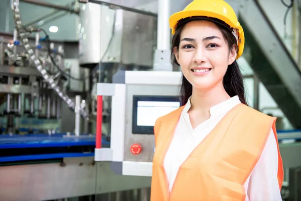 Young engineer inspecting production line. Young beautiful chinese woman in safety hat posing and smiling with bottle filling production line in background.