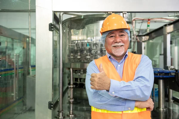 Senior engineer inspecting production line. Old chinese man in safety hat thumbs up with bottle filling production line in background.