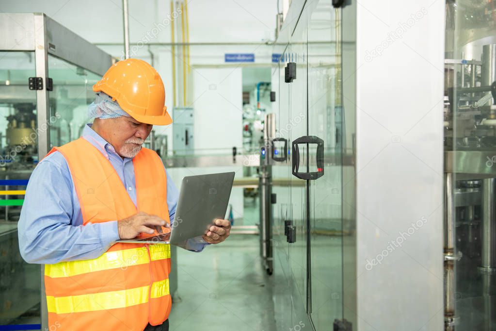 Senior engineer inspecting production line. Old chinese man in safety hat using laptop with bottle filling production line in background.