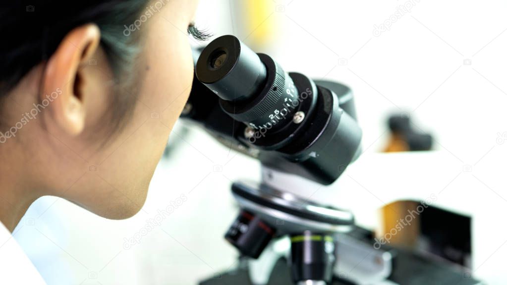 Young lab technician looking into mircoscope. Young beautiful chinese woman in lab coat looking into microscope with scient lab in background.