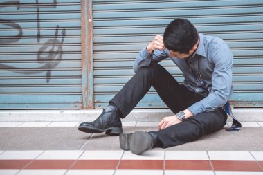 Sokakta oturan işsiz adam. Asya adam stres bakarak ve kapalı mağaza arka plan ile duygusal.