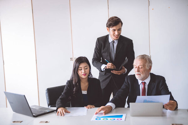 Старший бизнесмен обсуждает работу со своей командой. Старый кавказский мужчина в костюме с красивой молодой азиаткой, сидящей вместе, обсуждают бизнес-план. Концепция межпоколенческой бизнес-команды
.