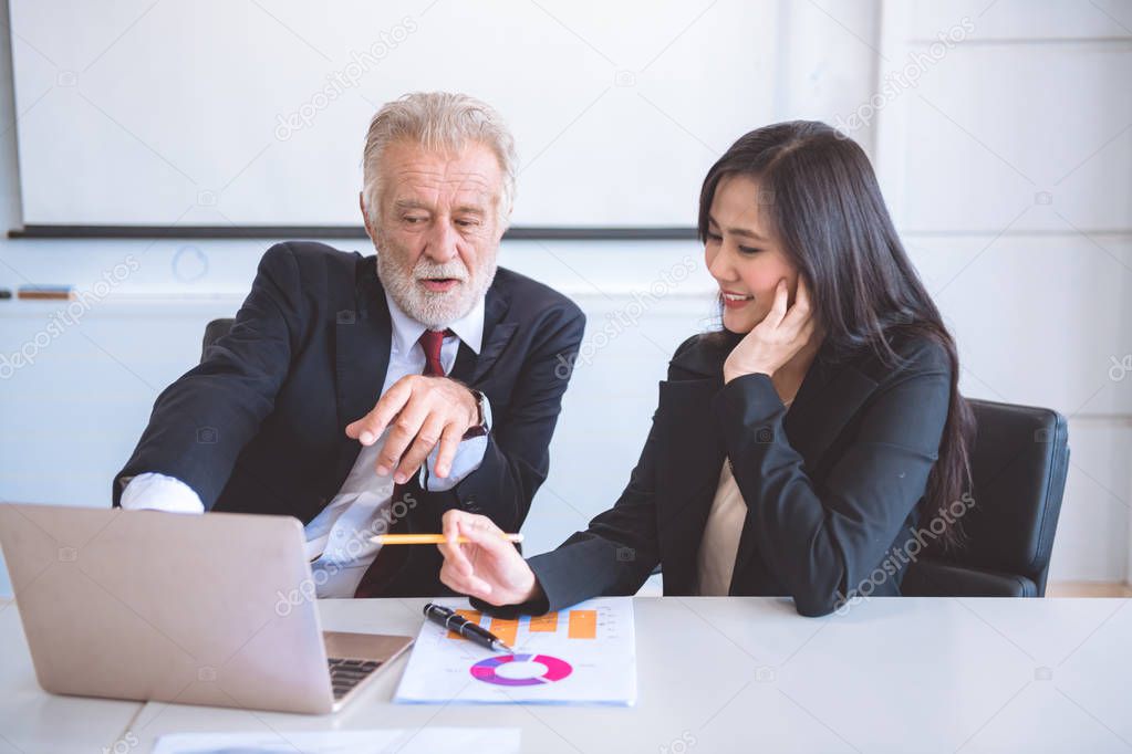 Senior businessman  pointing finger at laptop. Old caucasian male in suit with beautiful young asia woman sitting together discuss business plan. Cross generation business team concept.