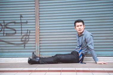 Sokakta oturan işsiz adam. Asya adam stres bakarak ve kapalı mağaza arka plan ile duygusal.