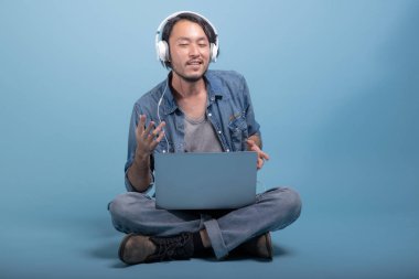 Genç adam oturup bilgisayarla mavi arka planda katta sakallı. Asya genç hippi müzik, tam vücut vurdu dizüstü kullanarak rahatla. Genç nesil yenilikçi çalışma kavramı.