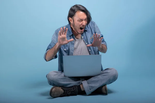 若いひげを生やした男は青い背景にコンピューターを使用して床に座る ラップトップ フルボディ ショットを使用してアジアの若いヒップを怖がっています 若い世代の流行に敏感な働くコンセプト — ストック写真