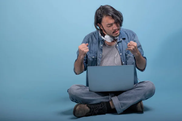 Young bearded man sit down on floor using computer in blue background. Disappointed Asian young hipster using laptop, full body shot. Young generation hipster working concept.