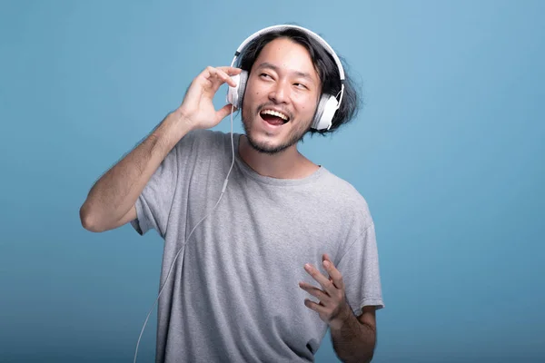 Young bearded man listening to music in blue background. Very Happy Asian young hipster using white headphone to listen to music, half body shot. Young generation hipster concept.