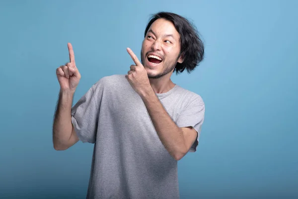 Young bearded man pointing finger in blue background. Excited Asian young hipster pointing finger at copy space, half body shot. Young generation hipster concept.