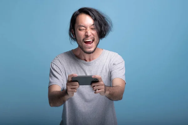 Excited young bearded man playing mobile games in blue background. Very happy Asian young hipster playing mobile game, half body shot. Young generation hipster relaxing concept.