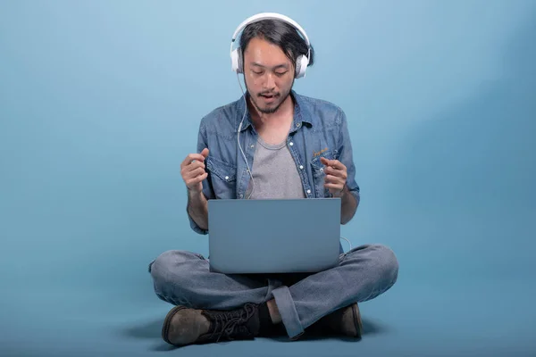 Young bearded man sit down on floor using computer in blue background. Relax Asian young hipster using laptop listening to music, full body shot. Young generation hipster working concept.