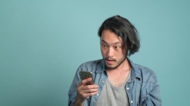 Genç erkek hipster yeni mobil ileti uygulamadan büyük bulmak için seyir onun telefon sakallı. Cep telefonu ve son derece mutlu o gördüğünüz gibi iyi haber mavi arka plan ile sıkılmış Asyalı adam.