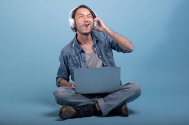 Genç adam oturup bilgisayarla mavi arka planda katta sakallı. Çok heyecanlı Asya genç hippi müzik, tam vücut vurdu dizüstü bilgisayar kullanarak. Genç nesil yenilikçi çalışma kavramı.