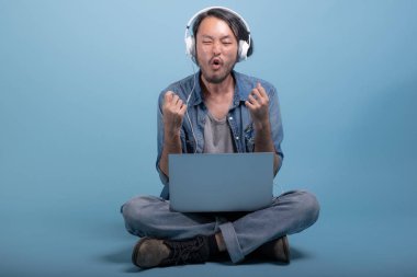 Genç adam oturup bilgisayarla mavi arka planda katta sakallı. Müzik, tam vücut vurdu dizüstü kullanarak heyecanlı Asya genç hippi. Genç nesil yenilikçi çalışma kavramı.