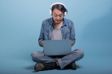 Genç adam oturup bilgisayarla mavi arka planda katta sakallı. Müzik, tam vücut vurdu dizüstü kullanarak endişeli Asya genç hippi. Genç nesil yenilikçi çalışma kavramı.