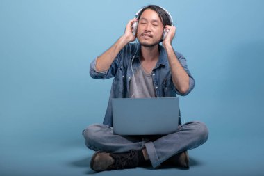 Genç adam oturup bilgisayarla mavi arka planda katta sakallı. Asya genç hippi müzik, tam vücut vurdu dizüstü kullanarak rahatla. Genç nesil yenilikçi çalışma kavramı.