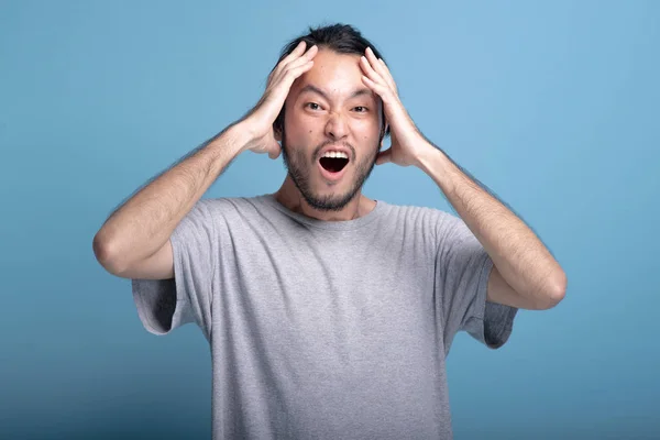Young bearded man excited shocking pose in blue background. Extremely surprise Asian young hipster with hands cover face, shocking expression, half body shot. Young generation hipster concept.