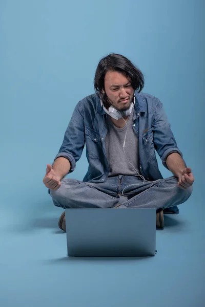 Young bearded man sit down on floor using computer in blue background. Confused Asian young hipster using laptop, full body shot. Young generation hipster working concept.