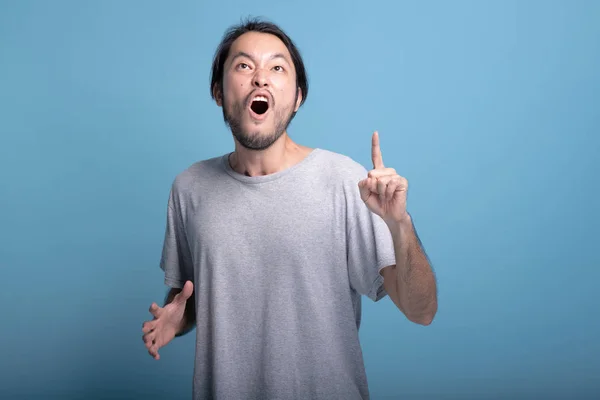 Young bearded man pointing finger in blue background. Extremely Excited Asian young hipster pointing finger upwards, half body shot. Young generation hipster concept.