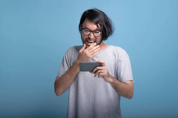 Upset young bearded man playing mobile games in blue background. Disappointed  Asian young hipster playing mobile game, half body shot. Young generation hipster concept.