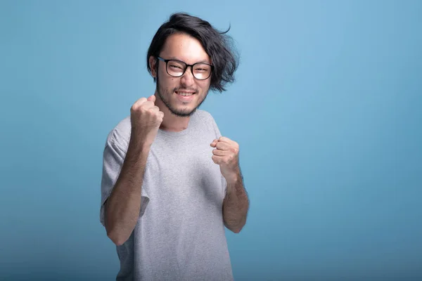 Young bearded man winning pose in blue background. Asian young hipster closed fist winner pose, half body shot. Young generation hipster concept.