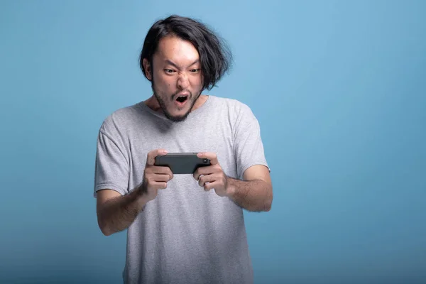 Excited young bearded man playing mobile games in blue background. Extremely excited Asian young hipster playing mobile game, half body shot. Young generation hipster relaxing concept.