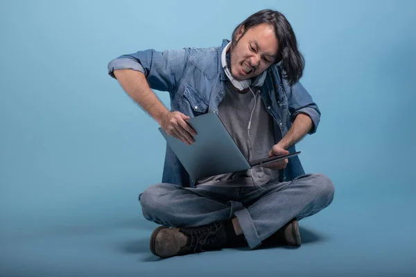 Young bearded man sit down on floor using computer in blue background. Very angry Asian young hipster using laptop, full body shot. Young generation hipster working concept.