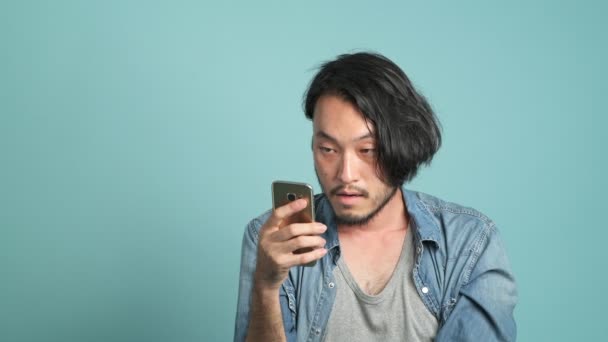 年轻的大胡子男性时髦看着他的手机 找到有趣的笑话从移动消息应用程序 无聊的亚洲人看着手机和笑大声 因为他看到有趣的笑话与蓝色背景 — 图库视频影像