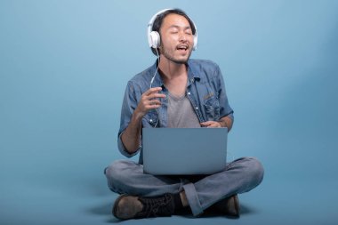 Genç adam oturup bilgisayarla mavi arka planda katta sakallı. Müzik, tam vücut vurdu dizüstü kullanarak çok mutlu Asya genç hippi. Genç nesil yenilikçi çalışma kavramı.