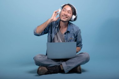 Genç adam oturup bilgisayarla mavi arka planda katta sakallı. Mutlu gülümseme Asya genç hippi müzik, tam vücut vurdu dizüstü bilgisayar kullanarak. Genç nesil yenilikçi çalışma kavramı.