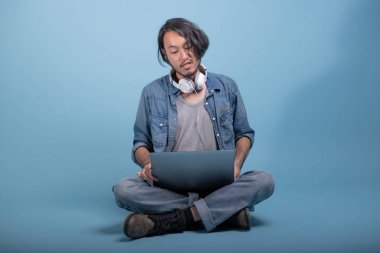 Genç adam oturup bilgisayarla mavi arka planda katta sakallı. Tam vücut vurdu, dizüstü bilgisayar kullanarak hayal kırıklığına Asya genç hippi. Genç nesil yenilikçi çalışma kavramı.