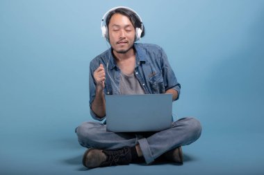 Genç adam oturup bilgisayarla mavi arka planda katta sakallı. Asya genç hippi müzik, tam vücut vurdu dizüstü kullanarak rahatla. Genç nesil yenilikçi çalışma kavramı.