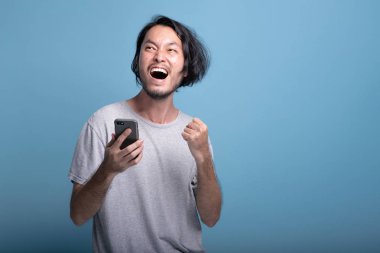 Heyecanlı genç adam arka plan mavi mobil ileti bakarak sakallı. Asya genç onun telefon, yarım vücut vurdu baktıktan sonra çok mutlu. Genç nesil yenilikçi kazanan kavramı.