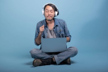 Genç adam oturup bilgisayarla mavi arka planda katta sakallı. Asya genç hippi müzik, tam vücut vurdu dizüstü kullanarak rahatla. Genç nesil yenilikçi çalışma kavramı.