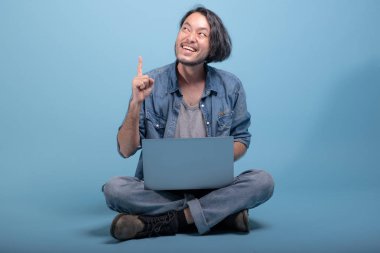 Genç adam oturup bilgisayarla mavi arka planda katta sakallı. Tam vücut vurdu, dizüstü bilgisayar kullanarak düşünme Asya genç hippi. Genç nesil yenilikçi çalışma kavramı.