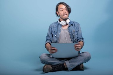 Genç adam oturup bilgisayarla mavi arka planda katta sakallı. Tam vücut vurdu, dizüstü bilgisayar kullanarak öfkeli Asya genç hippi. Genç nesil yenilikçi çalışma kavramı.