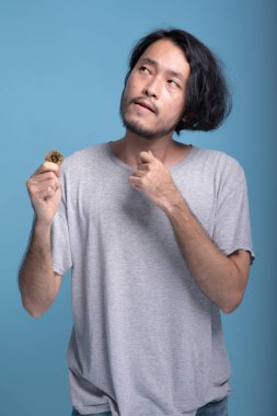 Genç bitcoin mavi arka planda tutan adam sakallı. Asya genç hipster holding altın bitcoin courious arıyorum düşünme, yarısı atış vücut. Genç nesil yenilikçi ticaret kavramı.