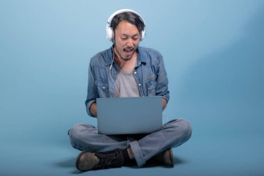 Genç adam oturup bilgisayarla mavi arka planda katta sakallı. Asya genç hippi müzik, tam vücut vurdu dizüstü kullanarak rahatla. Genç nesil yenilikçi çalışma kavramı.