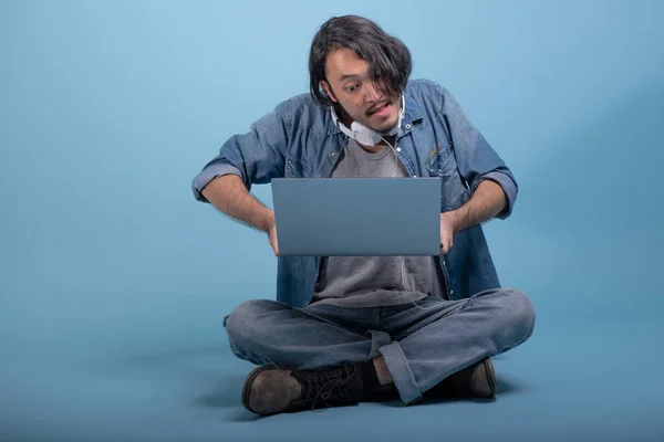 Young bearded man sit down on floor using computer in blue background. Angry Asian young hipster using laptop, full body shot. Young generation hipster working concept.