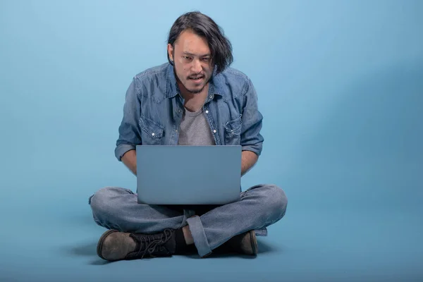 Young bearded man sit down on floor using computer in blue background. Focus Asian young hipster using laptop, full body shot. Young generation hipster working concept.