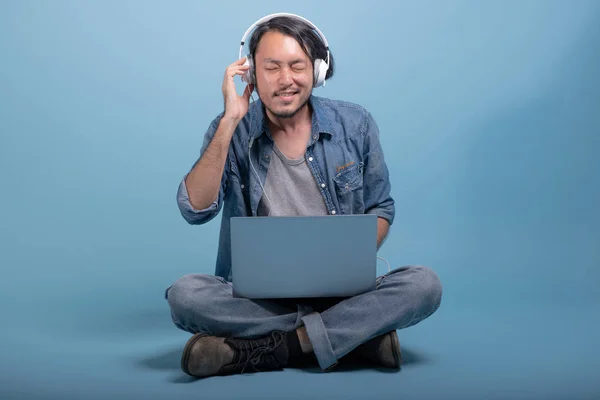 Young bearded man sit down on floor using computer in blue background. Relax Asian young hipster using laptop listening to music, full body shot. Young generation hipster working concept.