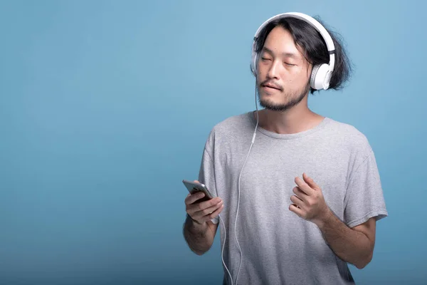 在蓝色背景下听音乐的年轻大胡子男子 放松亚洲年轻的时髦使用白色耳机听音乐 半身体射击 年轻一代时髦的概念 — 图库照片
